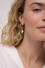 Close-up of a woman with wavy blonde hair wearing JaxKelly's Oval Pearl Drop earring, featuring 18k gold plating and a pearl. She is dressed in a white top with minimal makeup and a calm, relaxed expression.
