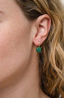 Close-up of a woman's ear wearing JaxKelly's Gemstone Mini Drop - Malachite earrings, featuring a rectangular green malachite gemstone in 18K gold plating. She has light skin and wavy blonde hair.