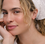 A woman with wavy blonde hair wears a white eyelet headscarf and JaxKelly Gold Long Oval Hoop earrings, resting her chin on her hand and smiling softly at the camera.