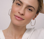 A woman with light skin and light brown hair pulled back smiles softly at the camera, wearing a white top and JaxKelly’s Silver Large Open Hoop earrings against a plain background.