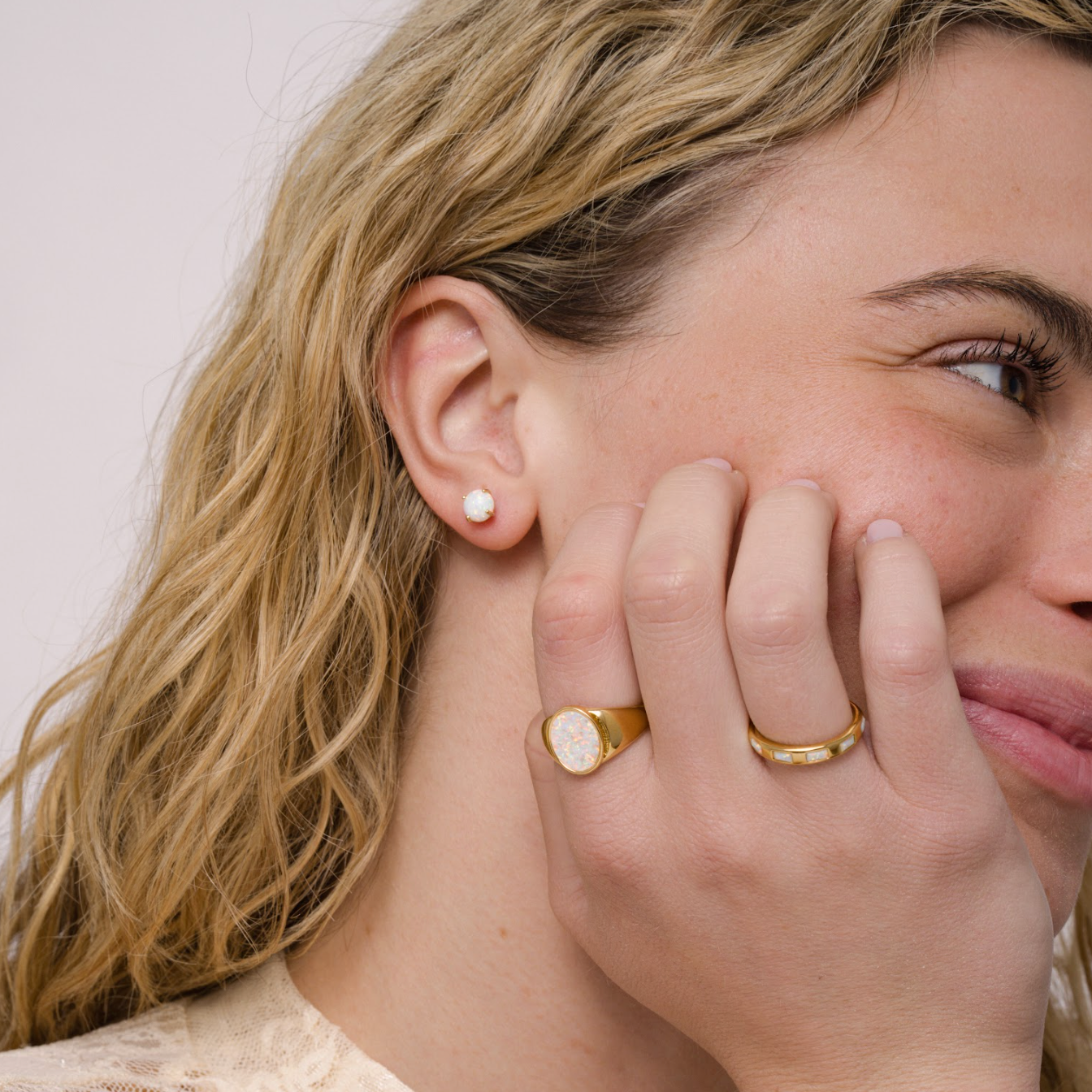 A woman with wavy blonde hair smiles softly, showing a pearl stud earring and two gold rings—one is the JaxKelly White Opal Signet Ring and the other 18k gold plated—on her hand near her face. She wears a light lace top with a subtle floral pattern.