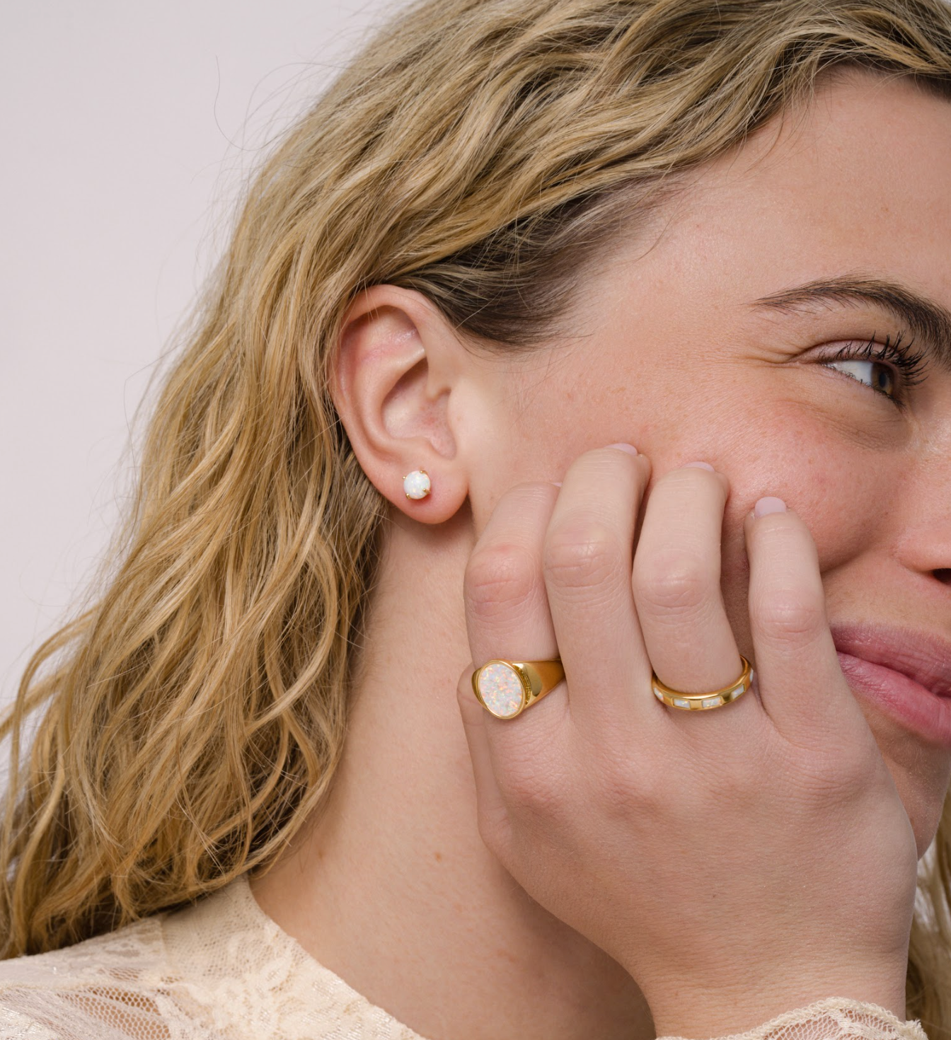 A woman with wavy blonde hair smiles softly, showing a pearl stud earring and two gold rings—one is the JaxKelly White Opal Signet Ring and the other 18k gold plated—on her hand near her face. She wears a light lace top with a subtle floral pattern.