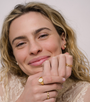 A smiling woman with wavy blonde hair in a lace top showcases the JaxKelly Chain Link Gold Ring, along with other gold jewelry, as she rests her chin on her hands against a plain background.