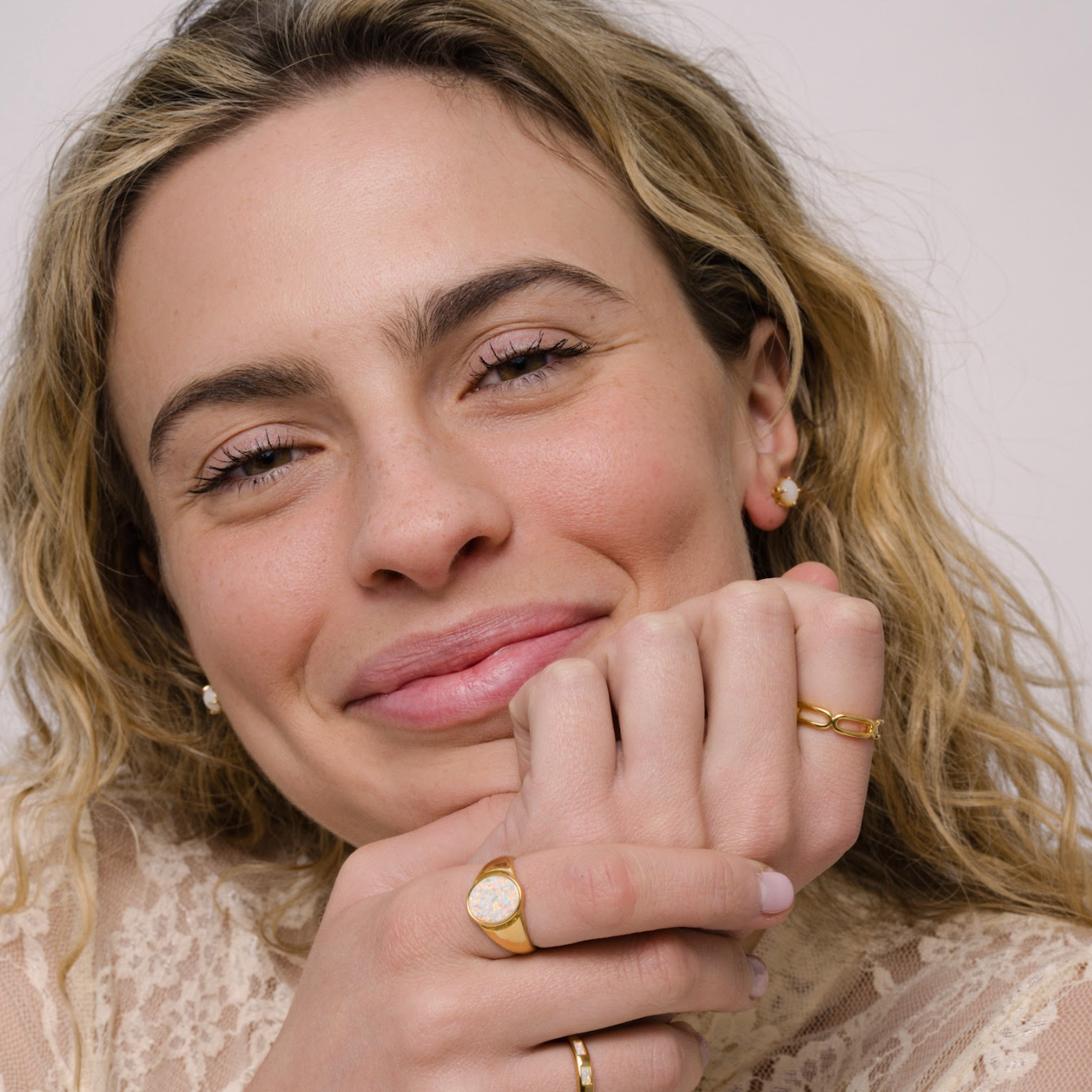 A smiling woman with wavy blonde hair in a lace top showcases the JaxKelly Chain Link Gold Ring, along with other gold jewelry, as she rests her chin on her hands against a plain background.