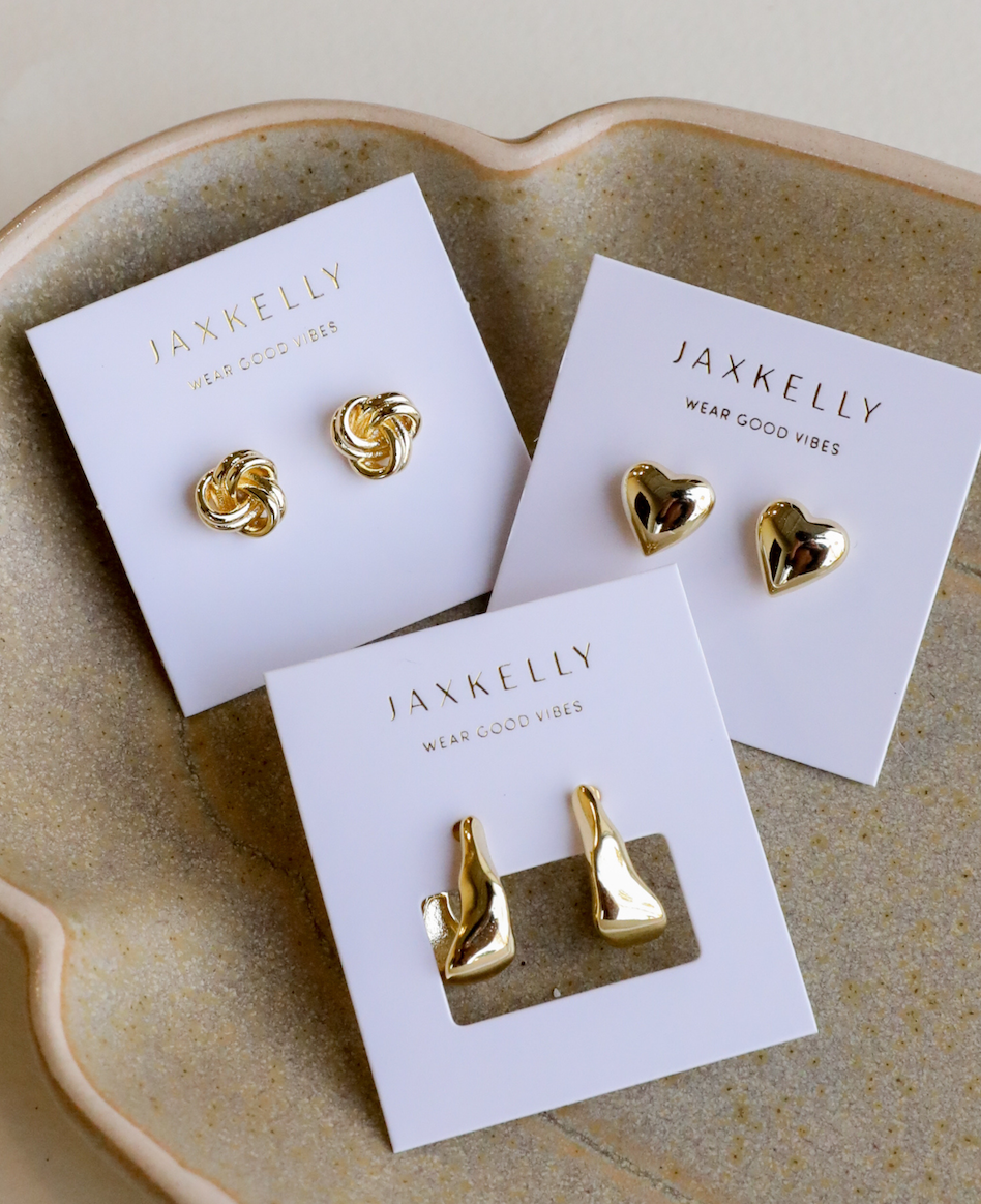 Three pairs of gold-plated earrings, including JaxKelly’s Puffed Gold Heart Earrings, knotted studs, and abstract drops, are displayed on white JaxKelly cards on a beige ceramic dish for a vintage-inspired touch.