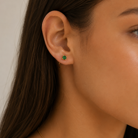 A close-up shows a woman wearing the JaxKelly Emerald CZ Huggie, a sparkling earring paired with a petite 18k gold plated hoop. Her long brown hair is tucked behind her ear, set against a simple light background.