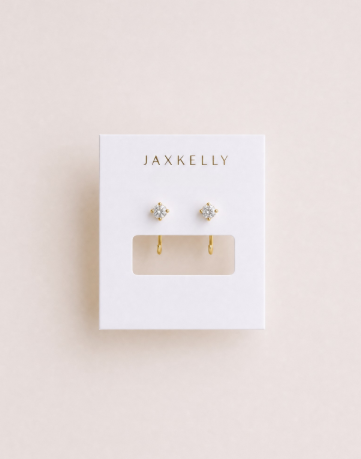A pair of small, tarnish-free gold White CZ Huggie earrings displayed on a white card with the brand name JaxKelly printed at the top.