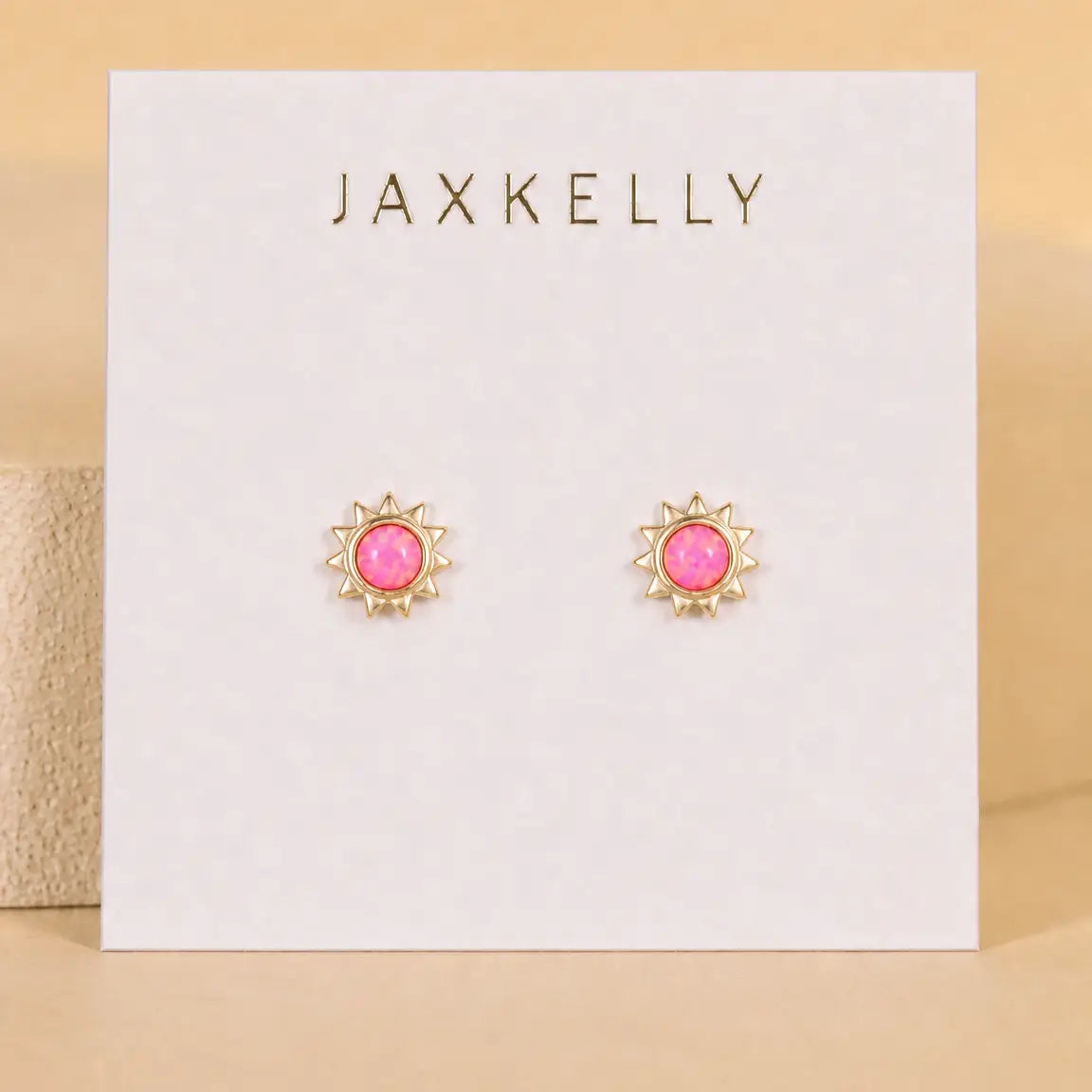 The Pink Opal Sun Stud by JaxKelly features gold sun-shaped studs with hypoallergenic posts and pink opal centers, presented on a white card stamped with the JaxKelly logo in gold.