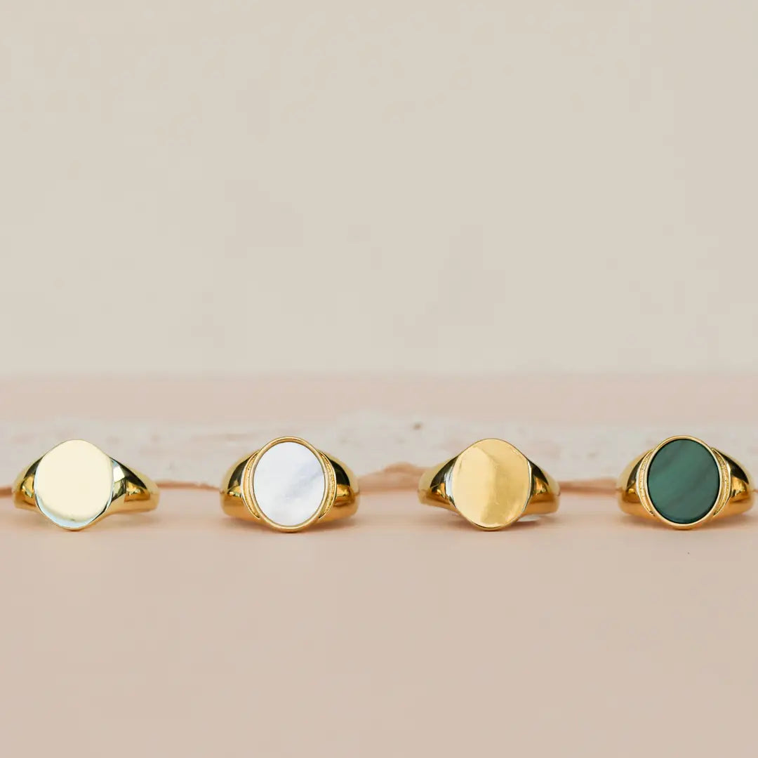 The JaxKelly Malachite Signet Ring is shown with three other 18k gold plated signet rings—each with unique stone inlays (green malachite, white, gold, and dark green)—lined up on a neutral surface against a soft beige background.