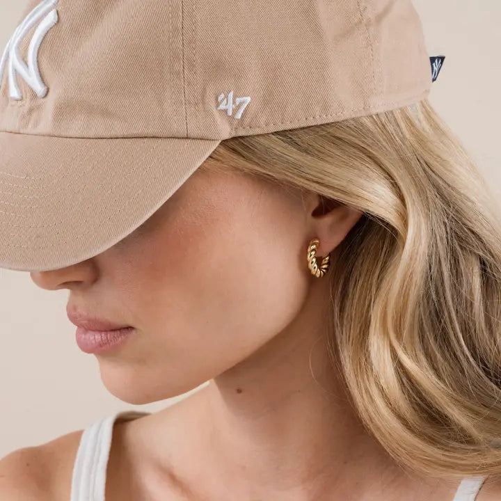 A woman in a beige New York Yankees cap and JaxKelly Small Channel Gold Hoop earrings with 18K gold plating, her wavy blonde hair loose, is shown in close-up side profile against a neutral background.