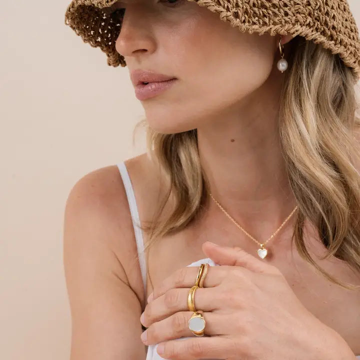 A woman in a white spaghetti strap top and wide-brimmed straw hat, with loose blonde hair, wears JaxKelly's Pearl Drop Earring and tarnish-free gold jewelry including rings and a heart-shaped necklace, gazing to the side.