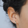 Close-up of an ear with short, curly dark hair wearing the Offset Trio - Amazonite: a small stud earring featuring three white amazonite stones on sterling silver posts and 18k gold plating. The background appears softly blurred.