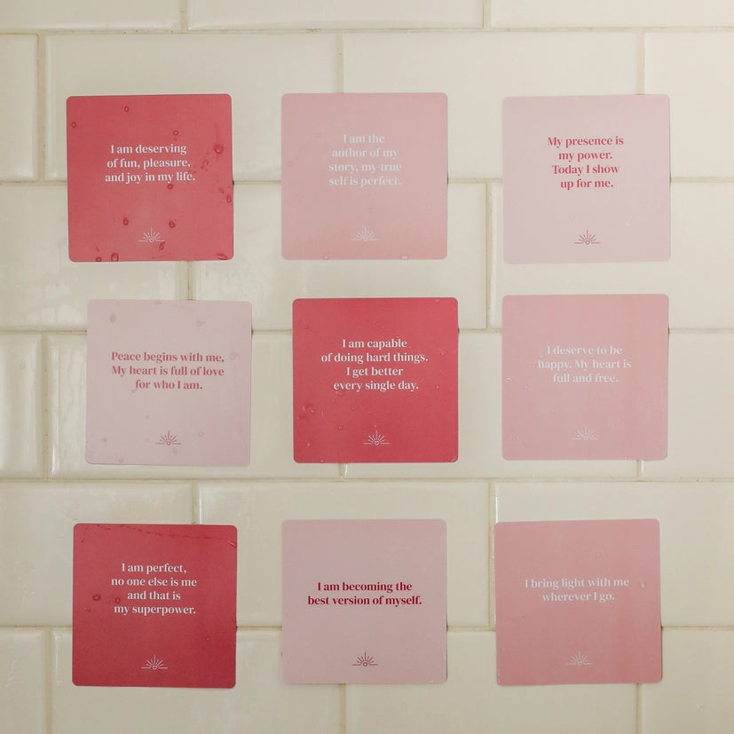 Nine Self-Love Shower Affirmations™ cards in pink, each waterproof and reusable, display unique self-love quotes in bold white text. The motivational cards are arranged in a 3x3 grid on a white tiled wall.