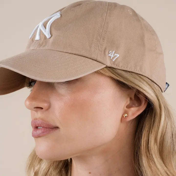 A woman with long blonde hair wears a beige baseball cap with a white NY logo and ’47 on the side. She accessorizes with JaxKelly’s Seashell Stud Earrings, which are hypoallergenic, tarnish-free, and heart-shaped, plus natural makeup.