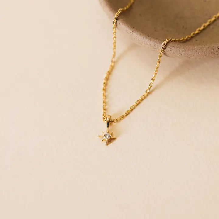 The JaxKelly Tiny Star Necklace, featuring an adjustable 18k gold plated chain with a small star-shaped Cubic Zirconia pendant, is shown on a neutral background beside a beige ceramic dish.