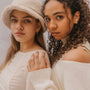 Two young women pose together in cream outfits. One wears a fuzzy bucket hat and JaxKelly Bold Hoop earrings, while her friend with curly hair dons an off-the-shoulder top. Both embody chic, confident style with lasting jewelry appeal.