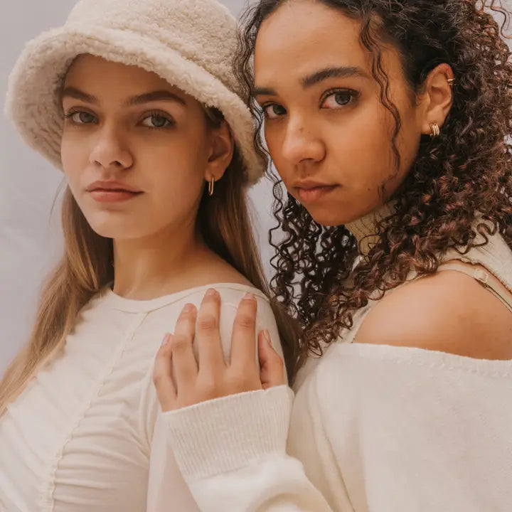 Two young women pose together in cream outfits. One wears a fuzzy bucket hat and JaxKelly Bold Hoop earrings, while her friend with curly hair dons an off-the-shoulder top. Both embody chic, confident style with lasting jewelry appeal.