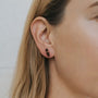 Close-up of a person’s ear wearing the JaxKelly Smoky Quartz Mineral Point - Protection earring. Their light brown hair is tucked behind the ear, and the neutral background is softly blurred.