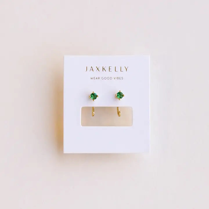 A pair of JaxKelly Emerald CZ Huggie earrings, 18k gold plated with small green stones, displayed on a white card featuring the WEAR GOOD VIBES slogan above the earrings.