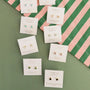 Nine pairs of JaxKelly Rose Quartz Mini Energy Gems - Love studs with 18k gold plating and sterling silver posts are displayed on labeled cards atop green and pink striped fabric, featuring gemstones like opal, turquoise, rose quartz, and amethyst.