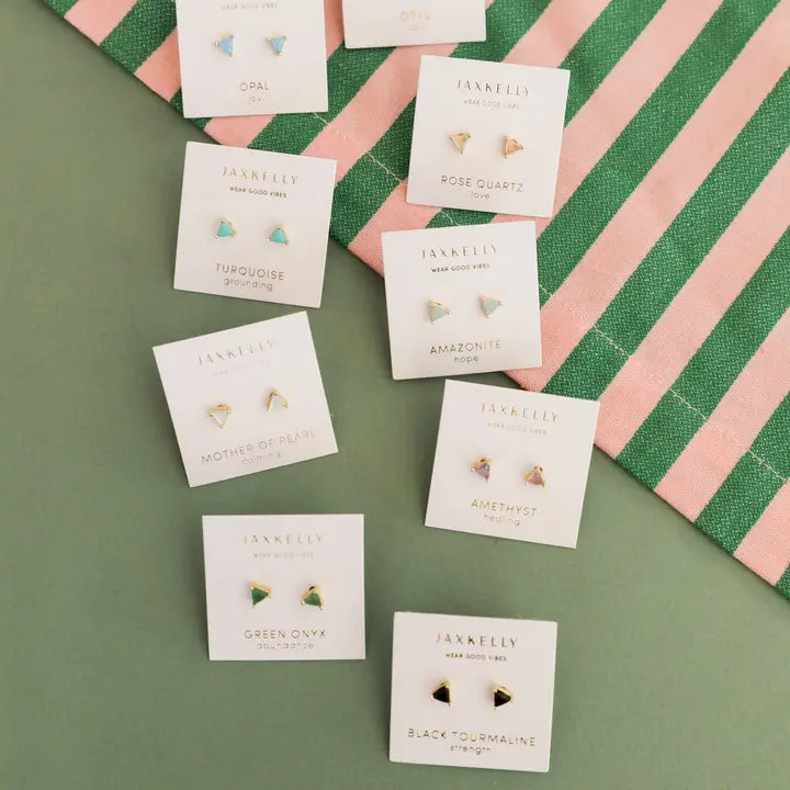 Nine pairs of JaxKelly Rose Quartz Mini Energy Gems - Love studs with 18k gold plating and sterling silver posts are displayed on labeled cards atop green and pink striped fabric, featuring gemstones like opal, turquoise, rose quartz, and amethyst.