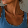 A woman in a denim sleeveless top wears the JaxKelly Gemstone Heart Pendant Necklace - Black Tourmaline, featuring a small heart-shaped pendant, shown from the shoulders up against a neutral background.