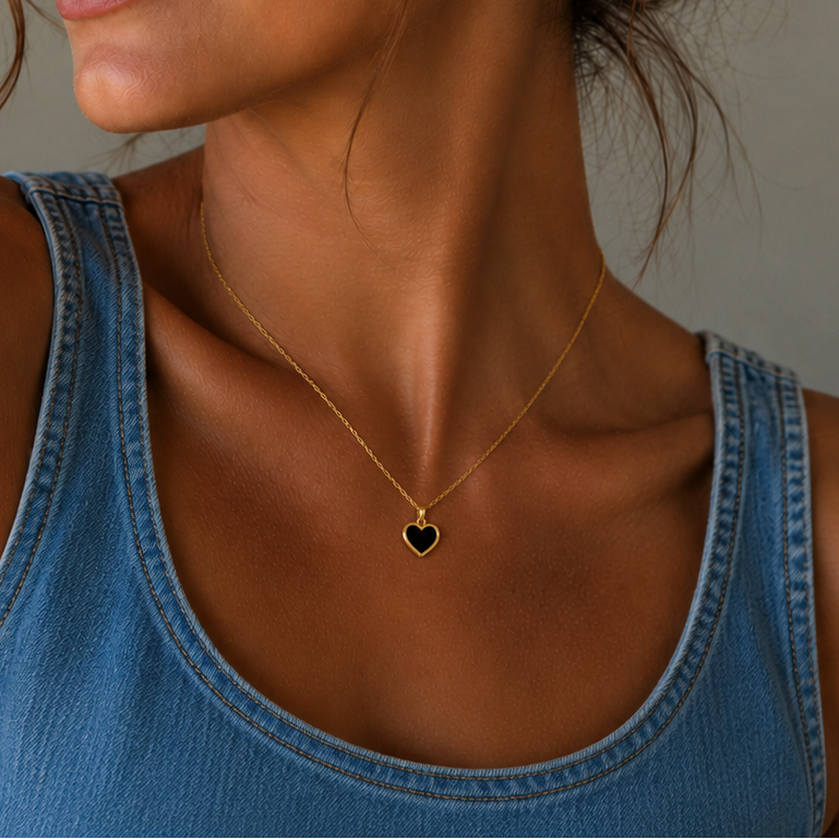 A woman in a denim sleeveless top wears the JaxKelly Gemstone Heart Pendant Necklace - Black Tourmaline, featuring a small heart-shaped pendant, shown from the shoulders up against a neutral background.