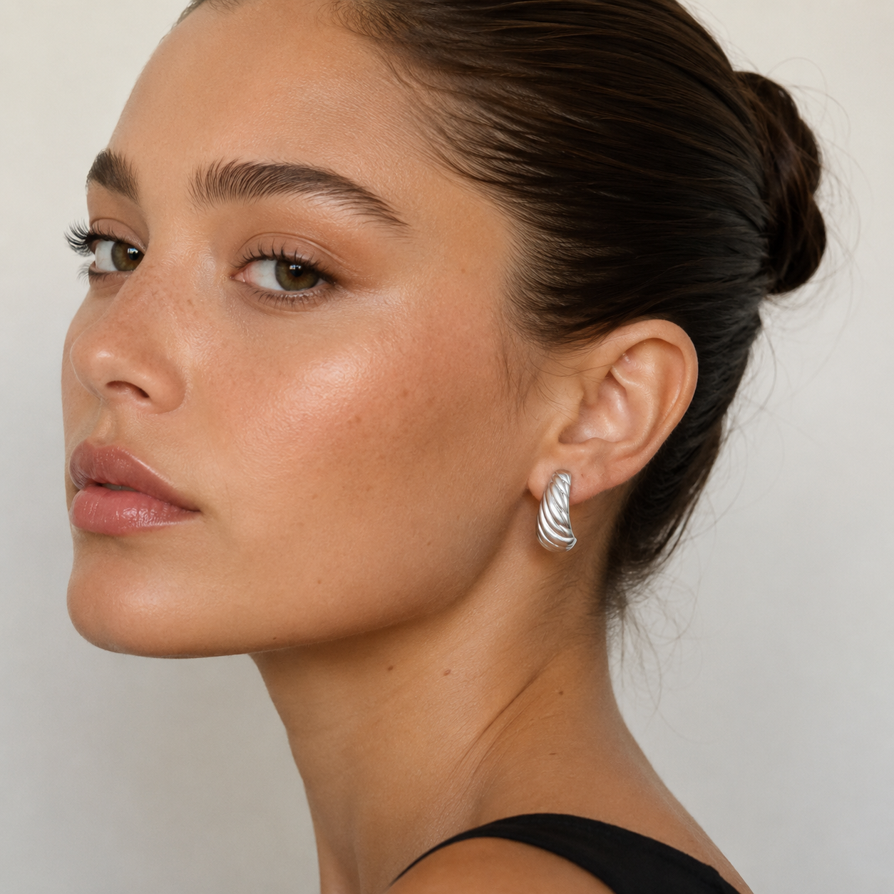 A woman with smooth, glowing skin and dark hair in a sleek bun wears a sleeveless black top and JaxKelly's Silver Bold Swirl Earrings, looking confidently at the camera against a neutral background.