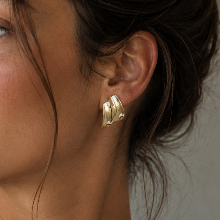 Close-up of a woman's ear wearing the JaxKelly Gold Bold Wave statement stud earring, featuring 18k gold plating and sterling silver posts. Her dark hair is loosely tied back, revealing smooth skin against a neutral backdrop.