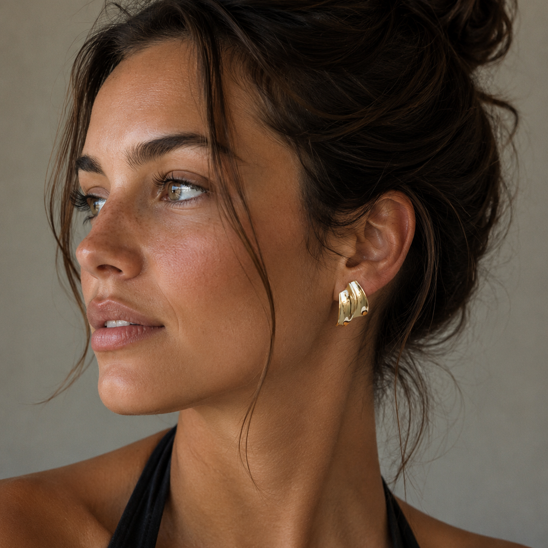 A woman with tan skin and brown hair in a loose updo wears the JaxKelly Gold Bold Wave chunky statement stud earrings with 18k gold plating and a black halter top, gazing to the side against a neutral background.