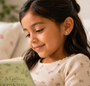 A young girl with long dark hair smiles while reading "A Gallery of Friends," wearing a cream, floral top and JaxKelly's Bookworm Enamel Flat Back Kids Earrings.