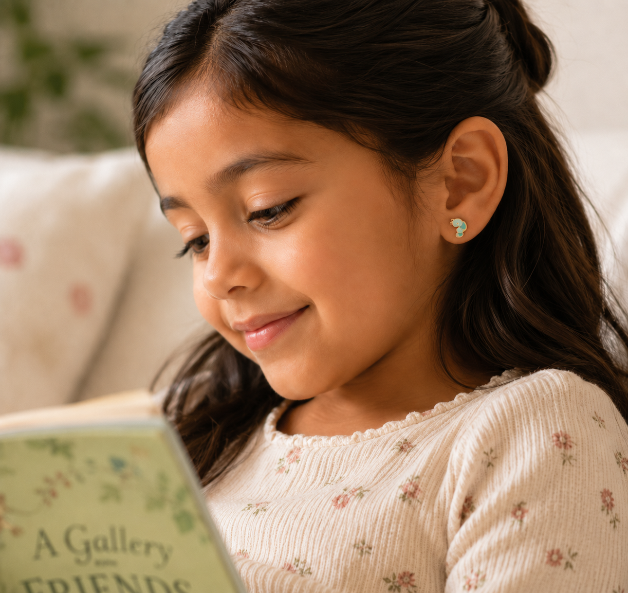 A young girl with long dark hair smiles while reading "A Gallery of Friends," wearing a cream, floral top and JaxKelly's Bookworm Enamel Flat Back Kids Earrings.