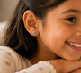 A young girl with long brown hair smiles, resting her chin on her hands. She wears a floral top and JaxKelly Bookworm Enamel Flat Back Kids Earrings—colorful, hypoallergenic, and perfect for sensitive ears. Only the right side of her face is visible.