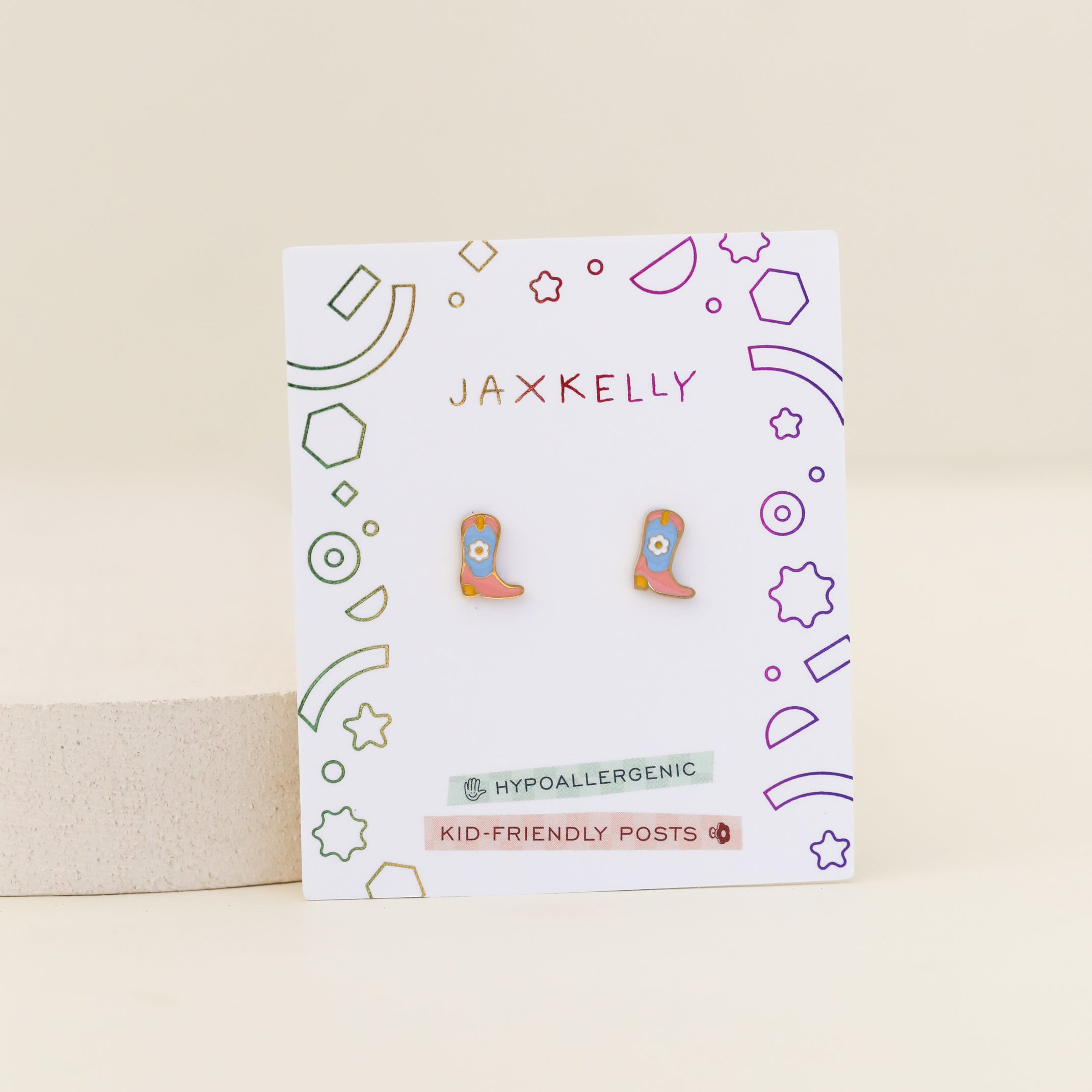 Boot Enamel Flat Back Kids Earrings in pink, shaped like cowboy boots with blue and yellow details. Displayed on a white JaxKelly card labeled "Hypoallergenic" and "Kid-friendly posts," with colorful geometric borders.