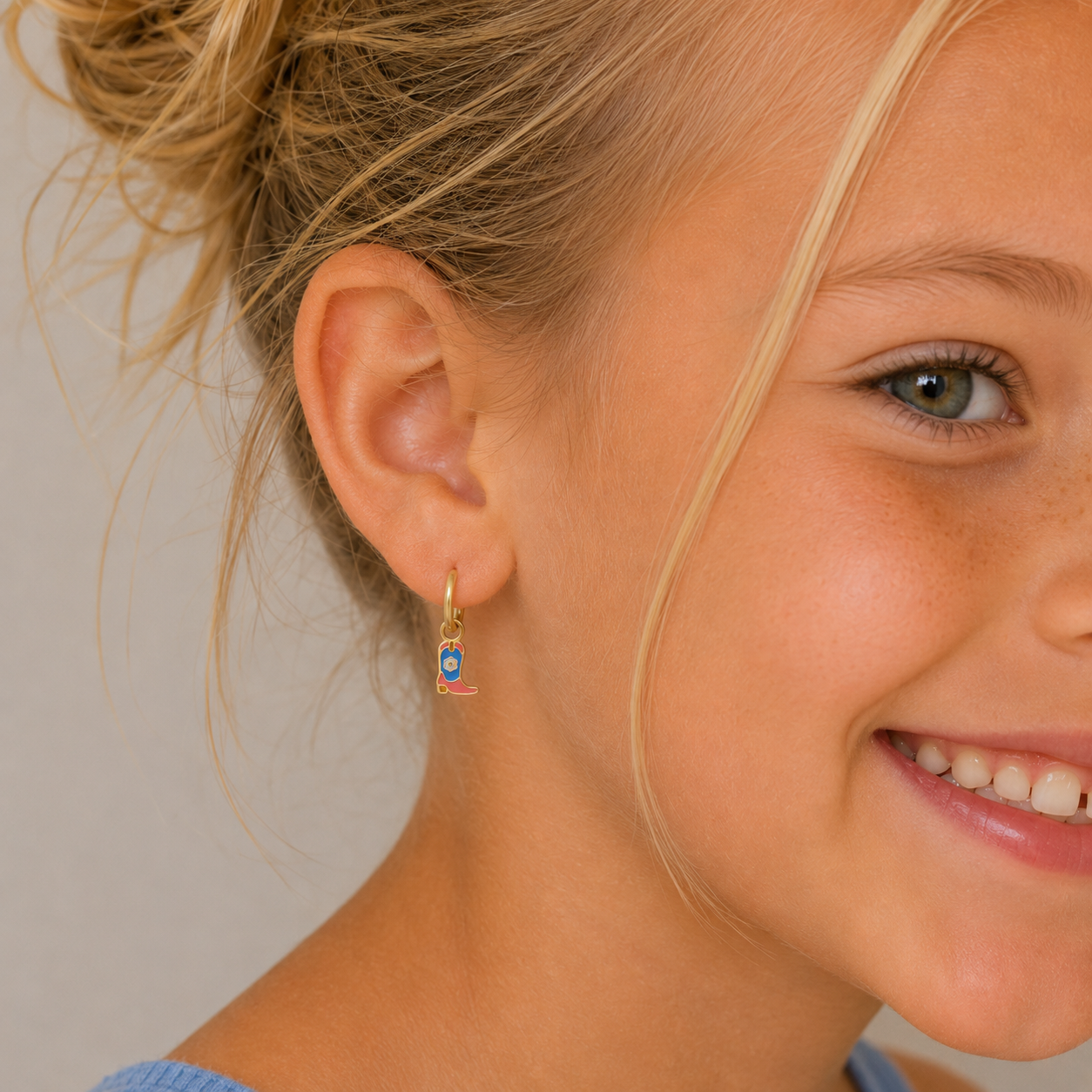 A young blonde girl in a light blue top smiles, showing off her JaxKelly Boot Enamel Charm Hoop—hypoallergenic kids earrings for sensitive ears with cute blue and pink charms. Only the right side of her face is visible.