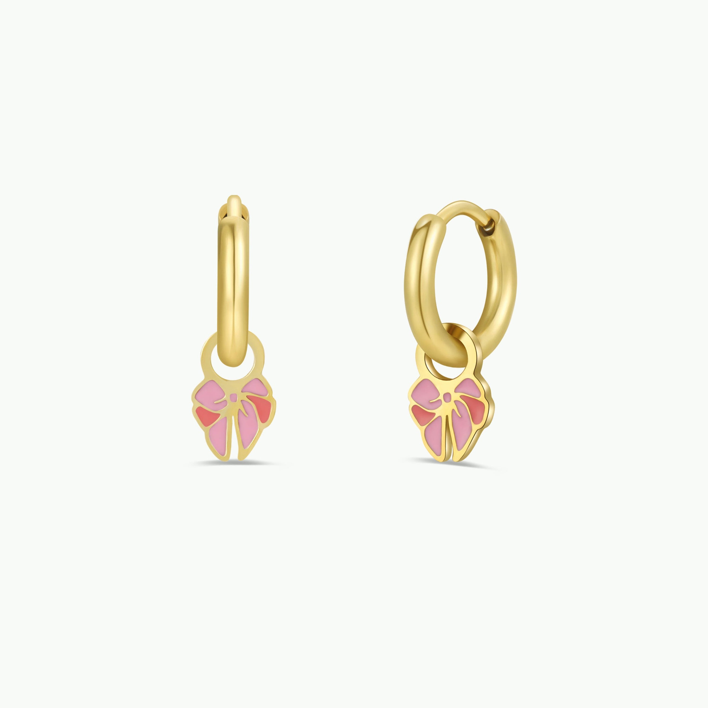 The Bow Enamel Charm Hoop features two small, hypoallergenic gold hoops, each with a pink and light gold enamel bow-shaped charm. These kids earrings are shown against a plain white background.