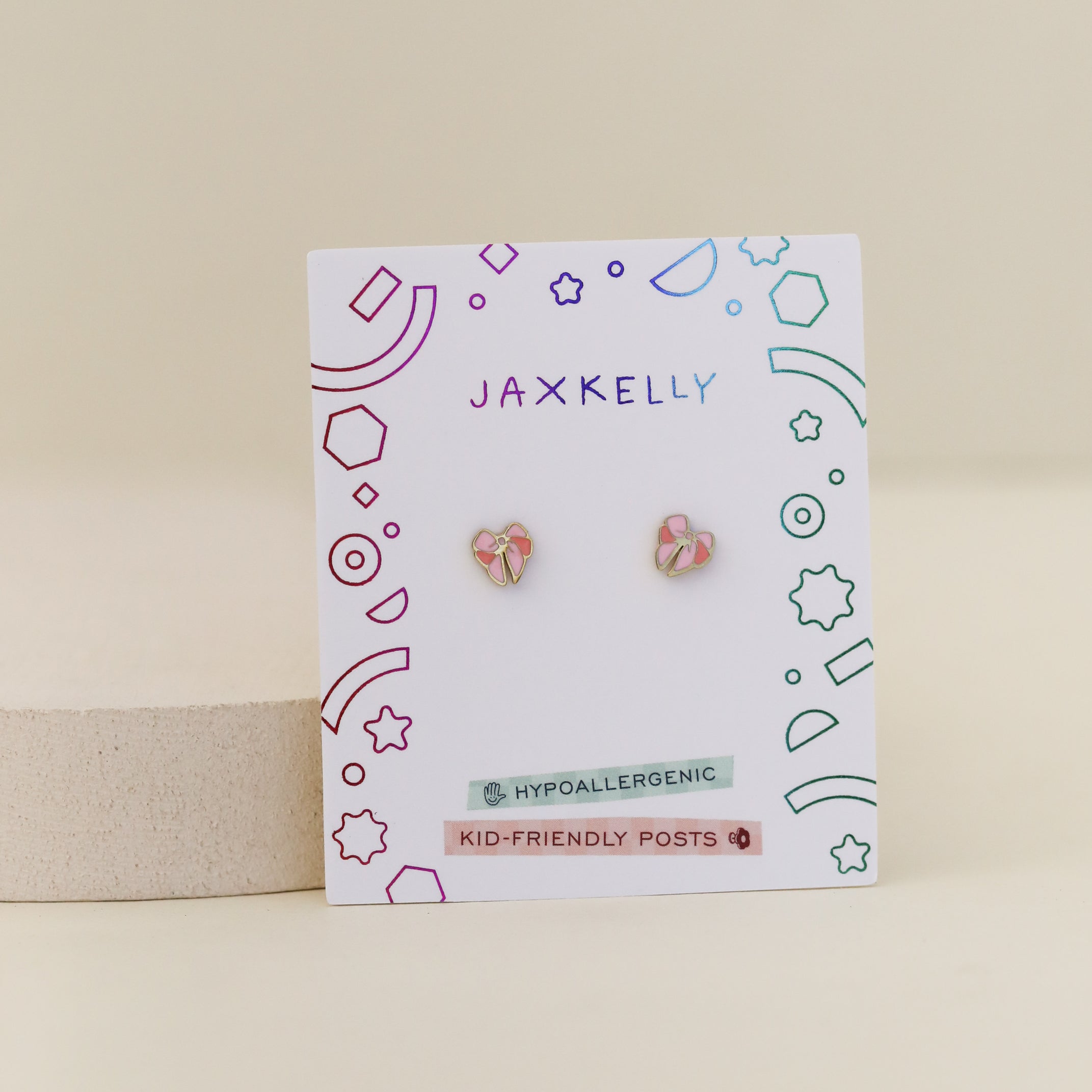 A pair of small, pink and gold Bow Enamel Flat Back Kids Earrings are shown on a white card with colorful geometric designs and the JaxKelly brand name. Labels indicate hypoallergenic, kid-friendly posts.