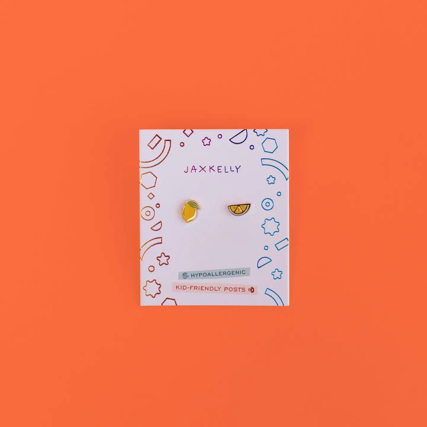 A pair of Lemon Enamel Flat Back Kids Earrings—one lemon slice and one orange slice—on a white JaxKelly card with colorful doodles, set against an orange background. Fun, playful and hypoallergenic!.