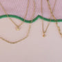Three JaxKelly necklaces—an 18k gold plated butterfly pendant, the Tiny Star Necklace with Cubic Zirconia, and an adjustable link chain—arranged on a pastel background with a wavy green embroidered border.