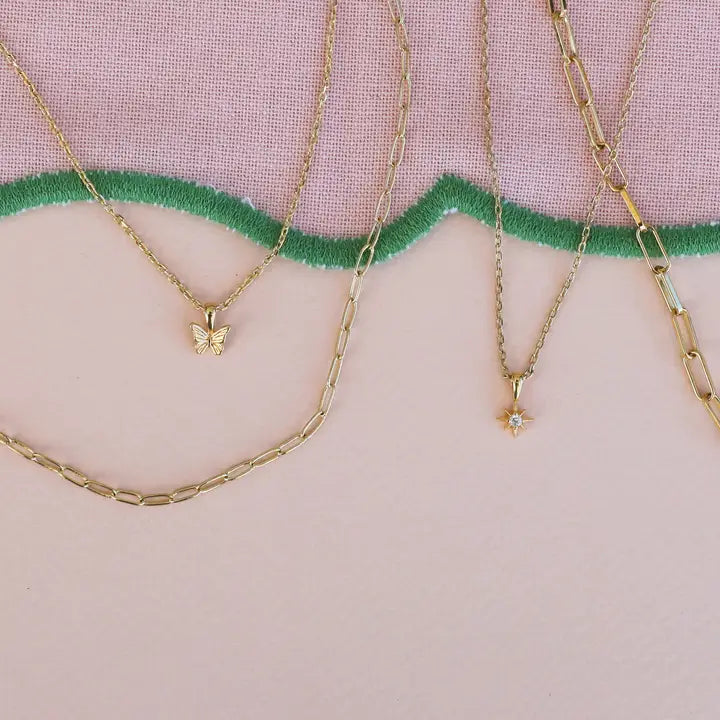 Three JaxKelly necklaces—an 18k gold plated butterfly pendant, the Tiny Star Necklace with Cubic Zirconia, and an adjustable link chain—arranged on a pastel background with a wavy green embroidered border.