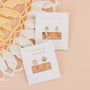 Two white “JaxKelly” cards display the brand’s Star Huggie earrings—gold hoops with starburst designs—resting on a light peach surface next to cream and yellow crocheted fabric.