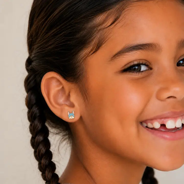A smiling young girl with dark braided hair wears JaxKelly Frenchie Enamel Flat Back Kids Earrings, facing the camera against a beige background.