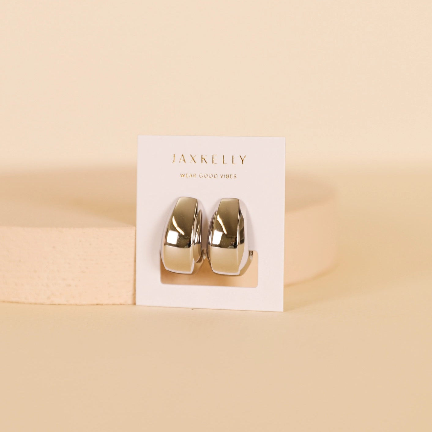 The Silver Bold Drop Earring features shiny, chunky statement hoops with sterling silver posts, displayed on a white JaxKelly card upright against a soft beige background.