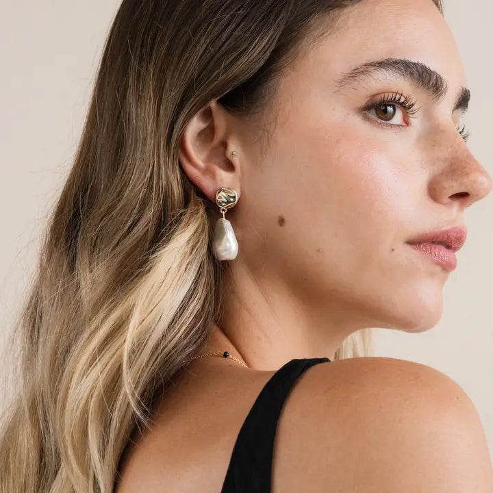 A woman with long, wavy, light brown hair wears JaxKelly Abstract Pearl and Gold Drop Earrings in hypoallergenic gold-plated metal. She has light skin, freckles, a black top, and is shown in profile against a neutral background.