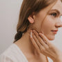 A young woman with light brown hair in a braid wears JaxKelly's Rose Quartz Hoop - Love earrings, featuring gold plating and a pink stone pendant. She gently touches her face, gazing sideways in a white top.