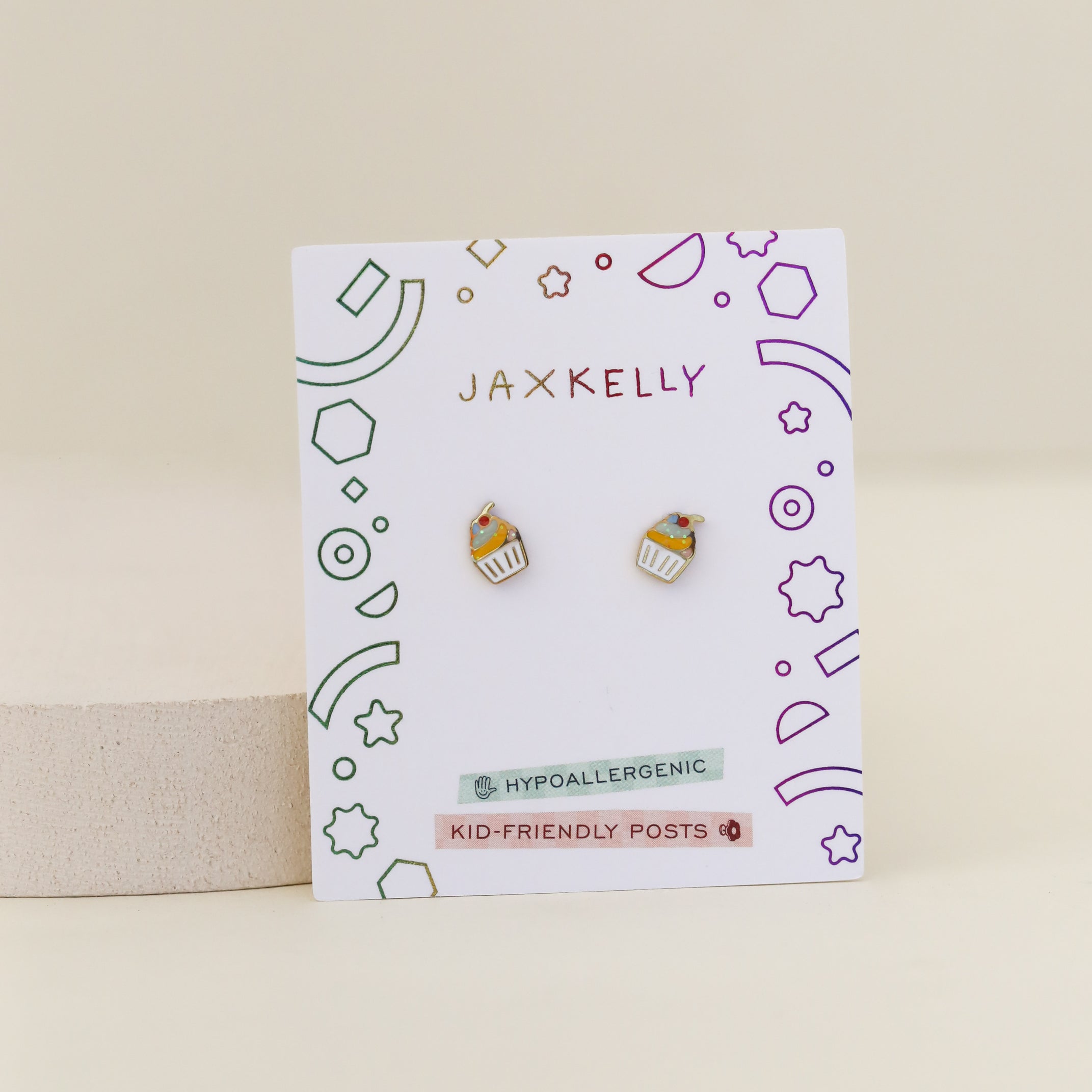 A pair of Cupcake Enamel Flat Back Kids Earrings on a JaxKelly card with colorful geometric designs. Hypoallergenic, these stud earrings are safe and comfortable for little ears.