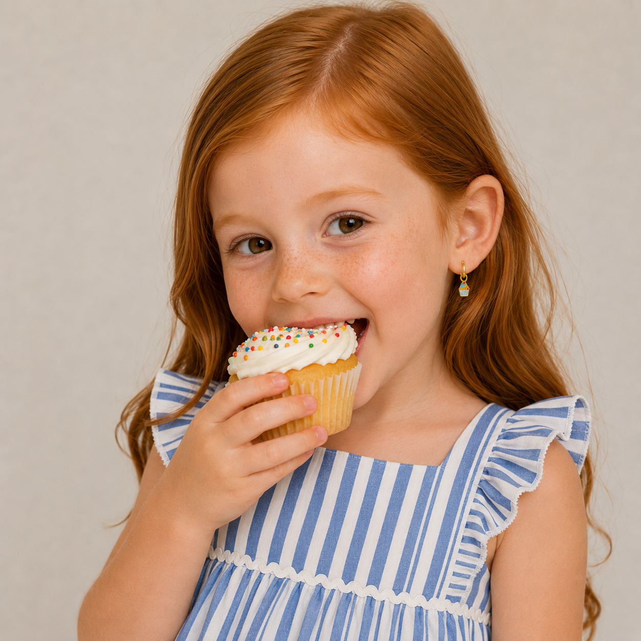 A young girl with long red hair in a blue and white striped dress smiles as she wears JaxKelly’s Cupcake Enamel Charm Hoop earrings, about to enjoy a cupcake topped with white frosting and colorful sprinkles.