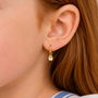 Close-up of a person with long auburn hair wearing the JaxKelly Cupcake Enamel Charm Hoop—a small, hypoallergenic gold earring shaped like a cupcake—designed for kids’ comfort; background and clothing appear out of focus.