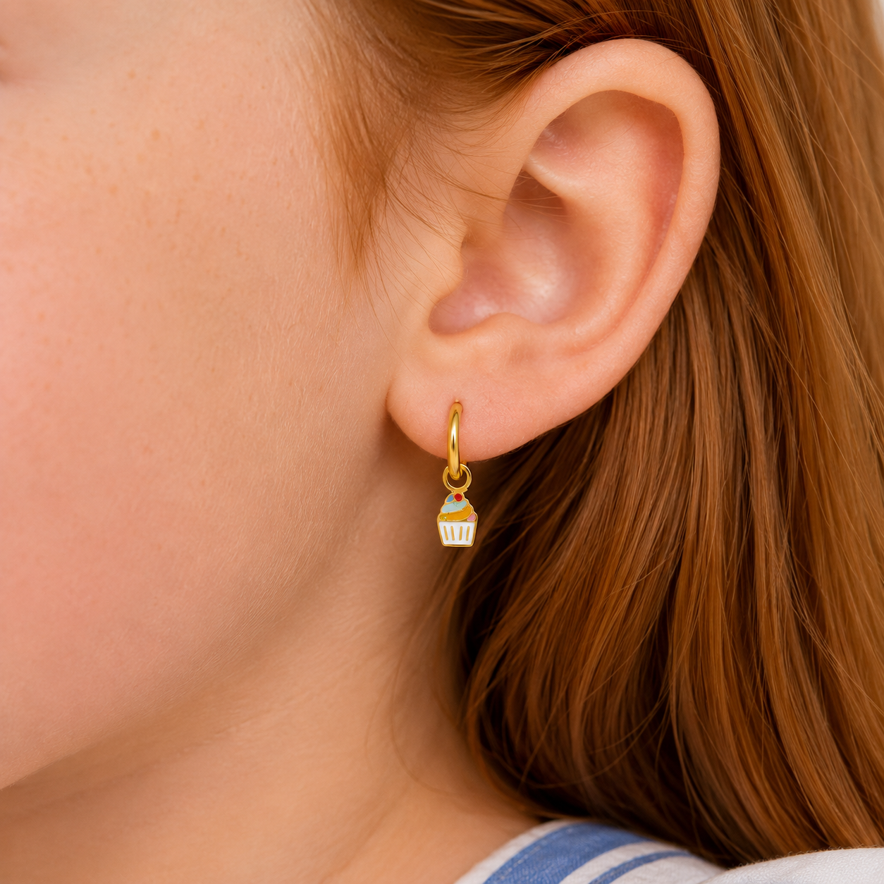 Close-up of a person with long auburn hair wearing the JaxKelly Cupcake Enamel Charm Hoop—a small, hypoallergenic gold earring shaped like a cupcake—designed for kids’ comfort; background and clothing appear out of focus.