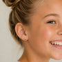 A smiling young girl with light brown hair in a bun wears a colorful Roller Skate Enamel Flat Back Kids Earring by JaxKelly in her left ear. Crafted from surgical-grade stainless steel, these hypoallergenic earrings are perfect for kids. Only the right side of her face is visible against a neutral background.